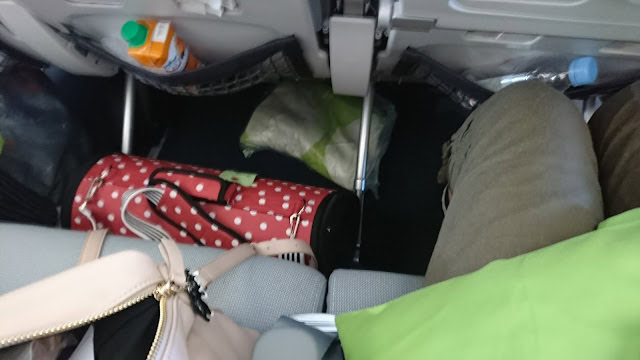飛行機内の座席と足元の荷物、隣の乗客の足元を写した写真です。.